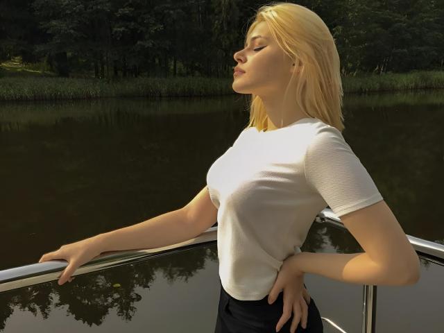 MissNestie, con cabello rubio, camiseta blanca y pantalones negros, de pie en un bote, con una mano en la barandilla, ojos cerrados, luz del sol en su rostro, reflejo en el agua..