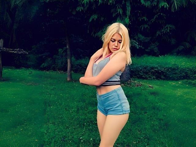 Blonde MissNestie with tits in a gray crop top and blue denim shorts, standing in a grassy park, arms crossed, looking down, in a sexy, confident pose.
