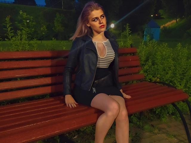 MissNestie, con cabello rosa, lápiz labial rojo, chaqueta de cuero negro, blusa a rayas, mostrando sus senos en una falda negra, sentada en un banco rojo por la noche, con las piernas cruzadas..