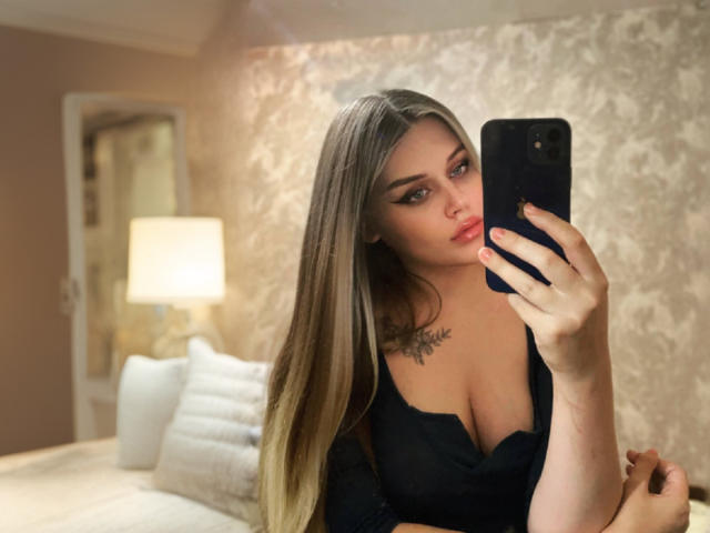 MissNestie with long blonde hair, wearing a low-cut black top, taking a selfie in a bedroom, showing her boobs and a tattoo on her chest. Holding a black phone.