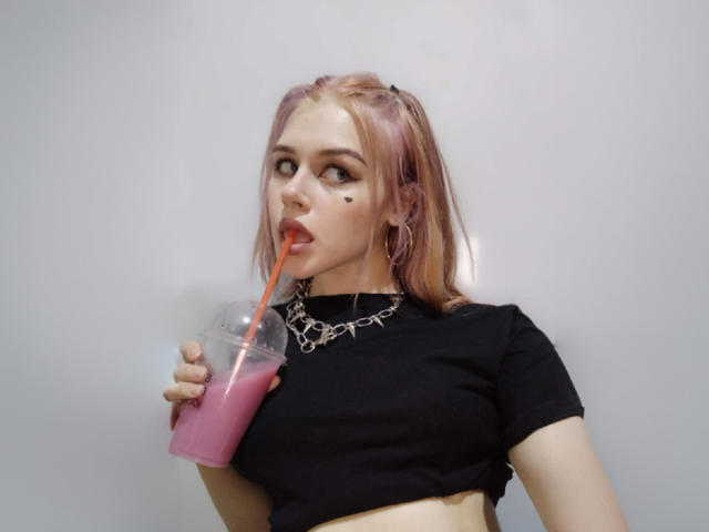 MissNestie with pink hair, black crop top, holding pink drink with straw, wearing spiked necklace, looking up, has black dot on cheek.