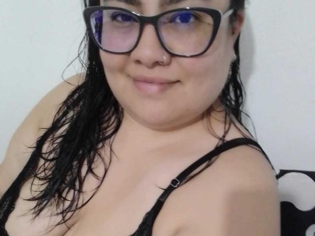 FoxyBell, avec ses cheveux noirs mouillés, ses lunettes, son anneau de nez et son soutien-gorge noir, affiche un léger sourire et ses seins nus de taille moyenne. Elle a la peau claire et un visage mignon et rond..