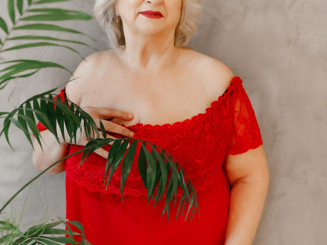 LolkaAlyss with pale skin, red lace off-shoulder dress, holding green palm leaves, gray short hair, red lipstick, touching her bare tits with one hand.