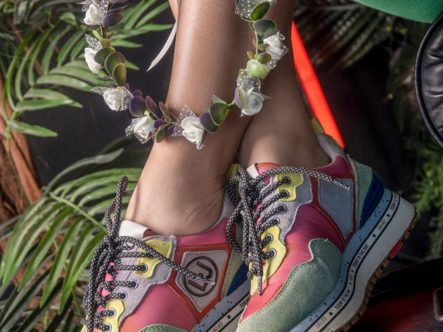 LettieSweet's legs in colorful, psychedelic sneakers with black, glittery laces, wearing a white and purple flower crown, showing her tan skin and slightly wrinkled ankles.