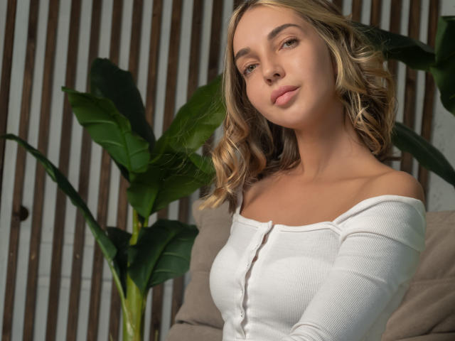 LettieSweet with blonde wavy hair, off-shoulder white ribbed shirt, small tits, light skin, and a subtle smile, standing in front of green plants and wooden slats.