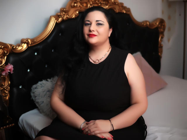 AleshaArabian hat eine blasse Haut, schwarzes, welliges Haar, roten Lippenstift, ein schwarzes Kleid, eine Kette um den Hals und sitzt auf einem schwarzen und goldenen, verzierten Bett, lächelnd, mit den Händen im Schoß, ein rosa Kissen im Rücken..