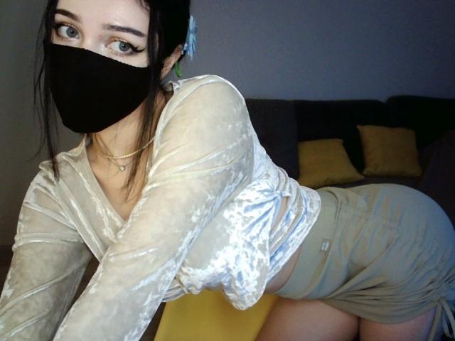 KathrineBerry with black mask, pale skin, blue eyes, and dark hair, wearing a sheer, white velvet top and tight, gray skirt, leaning forward, showing her ass.