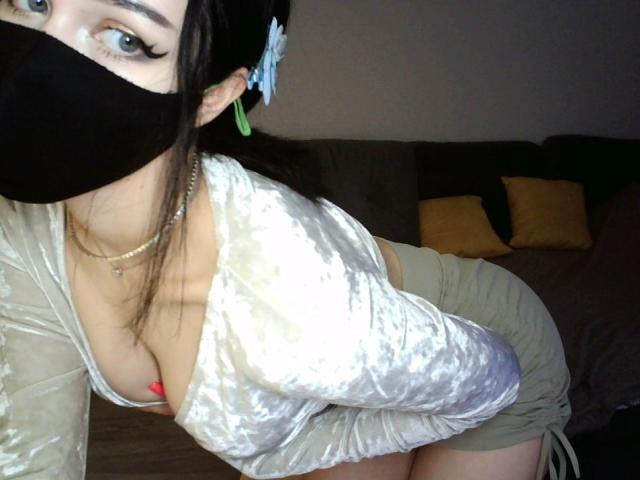 KathrineBerry with black mask, green earrings, white velvet top, showing her big tits and cleavage, green skirt, leaning forward, brown hair, green eyes, silver necklace, red bra visible.