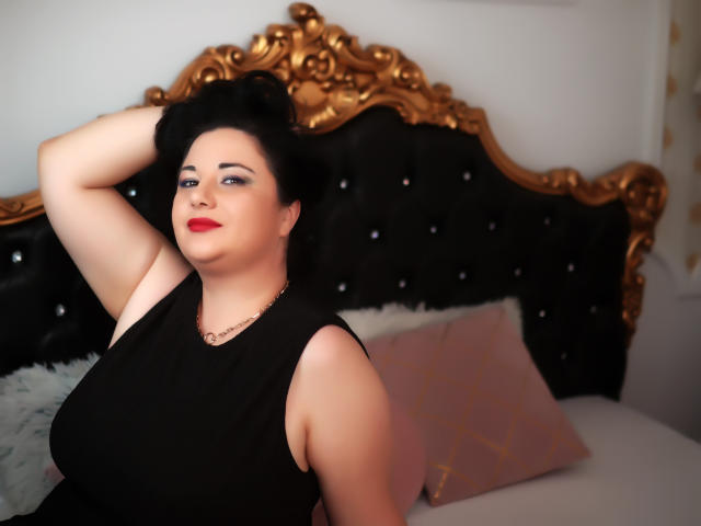 Alesha Arabian, formosa, con la pelle chiara e il rossetto rosso, indossa un abito nero senza maniche e ha un braccio alzato, seduta su un letto nero e dorato con imbottitura..