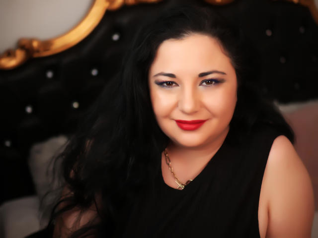 AleshaArabian with dark wavy hair, red lipstick, black sleeveless top, and gold necklace, smiling softly at the camera, showing her ass in the blurred background.