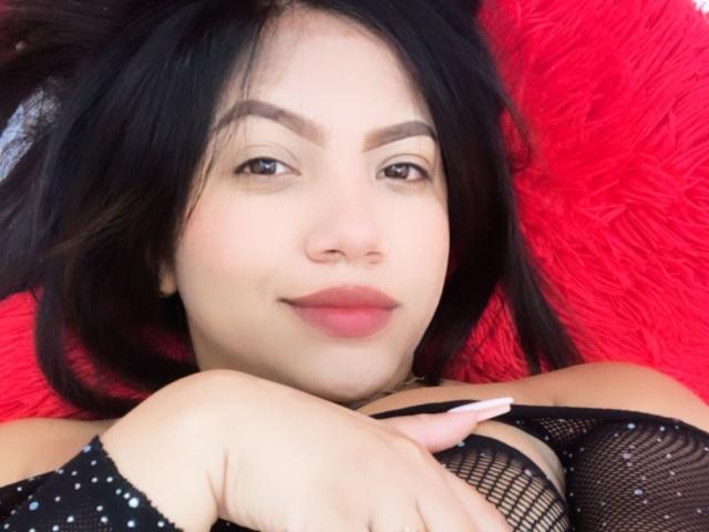 SashaCameron69, com cabelo preto, olhos castanhos, lábios vermelhos, vestindo uma blusa de rede preta, deitada em um travesseiro vermelho e fofo, com a mão no peito, sorrindo levemente, olhando para a câmera..