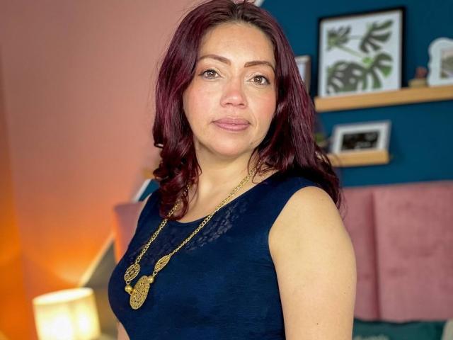 HelennaDuque, avec ses cheveux roux, porte un haut bleu foncé sans manches et un collier en or. Elle a une poitrine de taille moyenne et une expression légèrement boudeuse. Elle se tient dans une pièce très éclairée..