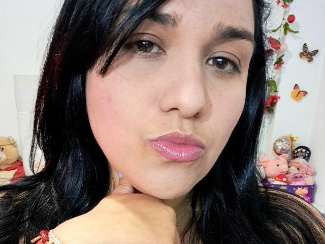 Viviana Thompson tiene cabello negro, ojos marrones, labios rosados y brillantes, y una mano en su barbilla. Está mirando directamente a la cámara con un ligero ceño fruncido. No se ven los senos..