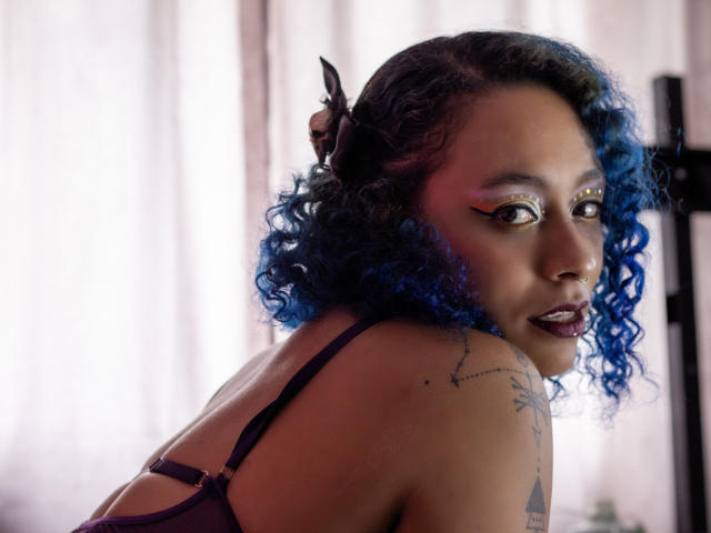 CataXBlue with blue curly hair, dark lipstick, and bold eyeliner, looking over her shoulder, showing a tattoo on her upper arm, wearing a purple bra.