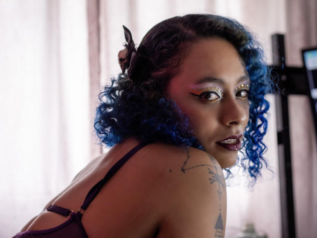 CataXBlue with blue curly hair, dark lipstick, and bold eye makeup, looking over her shoulder, showing a tattoo on her right shoulder. She's wearing a dark bra.