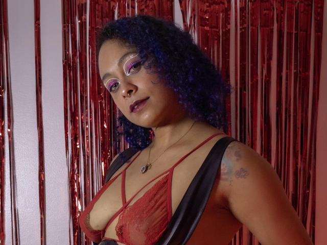CataXBlue with purple curly hair, red lace bra, black shawl, purple eyeshadow, dark lipstick, tattoos on right shoulder, necklace, red and silver background, looking at camera.