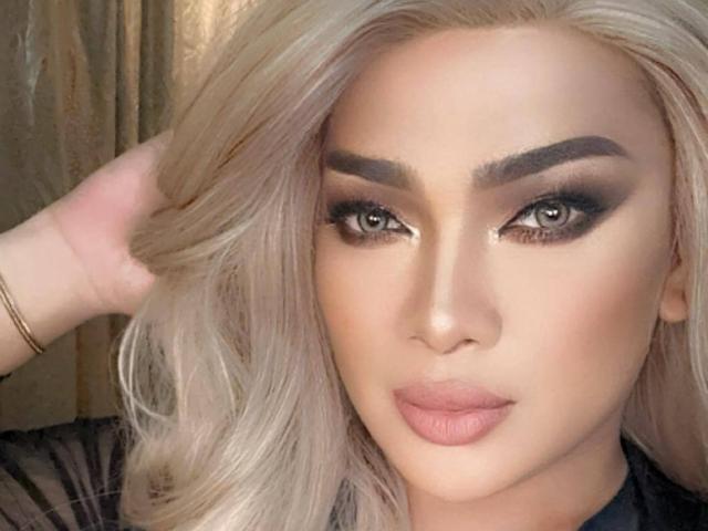 ScreenGodess has platinum blonde hair, thick eyebrows, smoky eye makeup, and full pink lips, with a hand in her hair, wearing a black sheer top, and a gold bracelet.