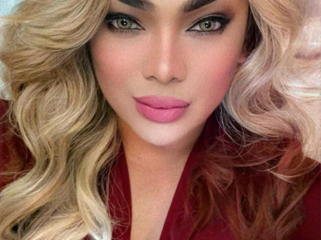 ScreenGodess a les cheveux blonds et ondulés, des yeux verts, des sourcils foncés, du rouge à lèvres rose, et porte un haut rouge. Elle regarde directement l'appareil photo avec une expression légèrement boudeuse..