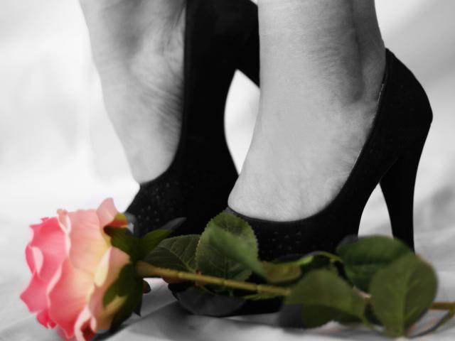 Les pieds de ChristinMoore, chaussés de talons hauts noirs, avec une rose rose en dessous, des feuilles vertes et un fond blanc. Les pétales roses de la rose ressortent sur les talons noirs..