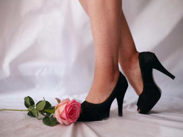 ChristinMoores Beine in schwarzen High Heels, mit einer rosa Rose dazwischen, liegen auf einem weißen Laken. Ihre Füße sind leicht gekreuzt, wodurch ihre nackte, glatte Haut sichtbar ist..