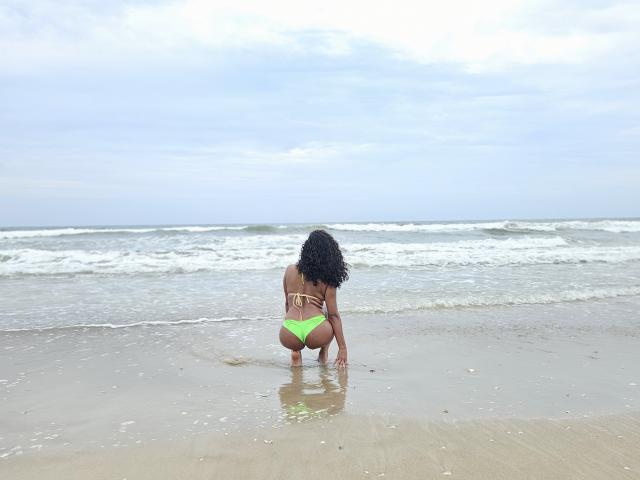 LolaPetterson with curly black hair, squatting on a beach, wearing a neon green thong bikini, showing her ass, facing the ocean, with gentle waves and cloudy sky.