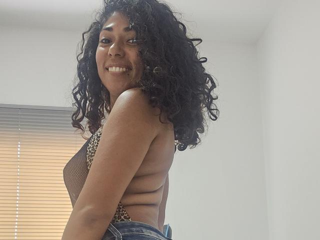 LolaPetterson with curly black hair, smiling, wearing a leopard print mesh top, showing her sideboob and back, in blue jeans, standing in a bright room.
