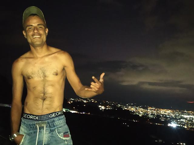 StrongAlan è senza camicia, ha un petto magro e peloso, e indossa jeans blu con una cintura bianca, indicando con un sorriso, e indossa un cappello verde di notte..