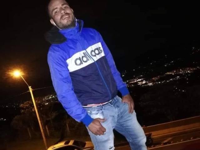 StrongAlan, een kale, baardgroeiige man in een blauwe Adidas jas en versleten jeans, staat 's nachts, glimlachend, met zijn handen in zijn zakken en met oordopjes in. Op de achtergrond staat een auto..