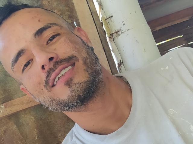 StrongAlan with a light brown beard and short hair, smiling with his tongue slightly out, wearing a white shirt, has a slightly dirty face and a relaxed expression.