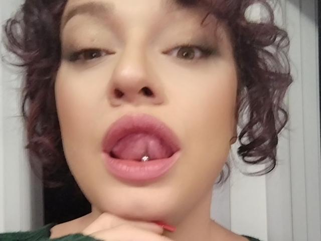 AliaBellia met krullend paars haar, licht open mond, tong met een klein piercing, en een groene top, met haar kin op haar hand rustend, waarbij haar roze lippen en bruine ogen zichtbaar zijn..