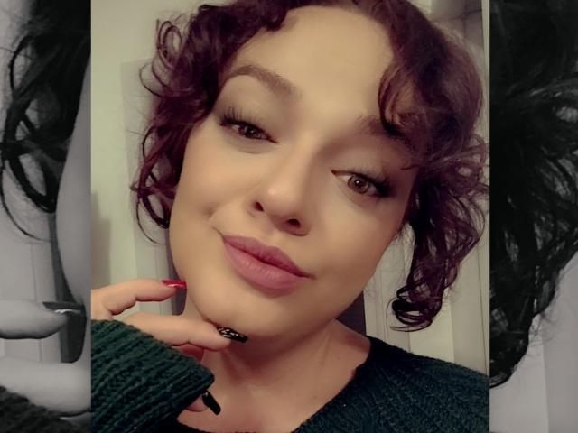 AliaBellia heeft krullend paars haar, roze lippenstift en een groene trui, met een schattige glimlach en een opgetrokken hand die zwarte en rode nagellak laat zien. Haar gezicht is gecentreerd..