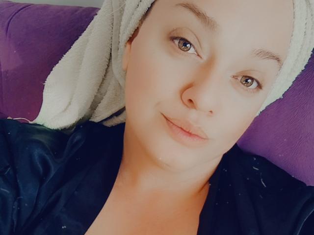 AliaBellia has light skin, brown eyes, and a towel on her head, wearing a black shirt, with a subtle smile, lying on a purple surface.