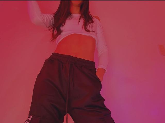 Lulu Jones lleva una camiseta blanca de manga larga y corte corto, que deja ver su abdomen, y pantalones negros holgados con una mano en el bolsillo, todo ello bajo una iluminación rosa y roja..