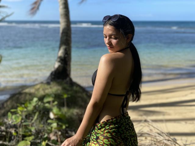 IssaPuasini with long black hair, wearing a black halter top and green patterned sarong, standing on a beach, looking back, sunglasses on head, palm tree in background.