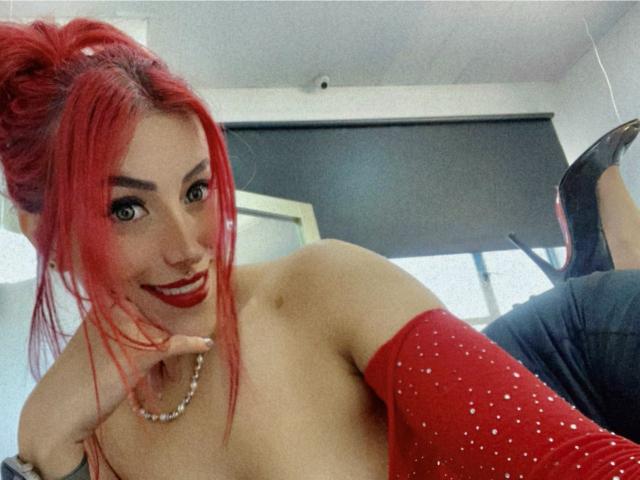 AlissaBrown with bright red hair, red lipstick, and red sparkly arm sleeve, smiling at the camera, showing off her bare tits, wearing a pearl necklace, in a mirror selfie.
