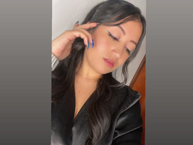 ShannelTaylor with long black hair, wearing a black velvet robe, has blue nails, closed eyes, and pink lipstick, touching her ear. Soft light highlights her smooth, fair skin.