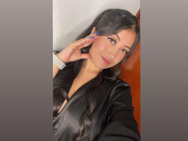 ShannelTaylor with long black hair, wearing a black satin shirt, blue nails, white wristband, smiling at camera, right hand touching ear.