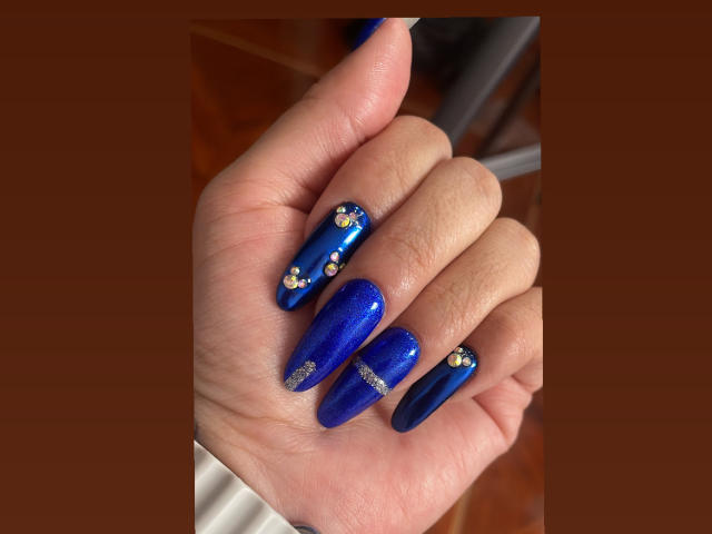 ShannelTaylor's hand with shiny blue nails, glitter accents, and small gold gems on the tips, against a brown background. Her nails are long and polished, looking sweet.