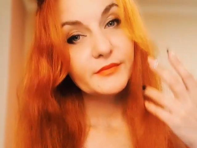 JasminsLove has orange-red hair, light skin, blue eyes, and orange lipstick. She's looking at the camera with a slight smile, touching her hair with black-nailed fingers.