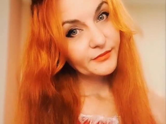 JasminsLove heeft feloranje haar, blauwe ogen en een schattige glimlach met oranje lippenstift. Ze kijkt naar de camera met een lichte kanteling van haar hoofd..