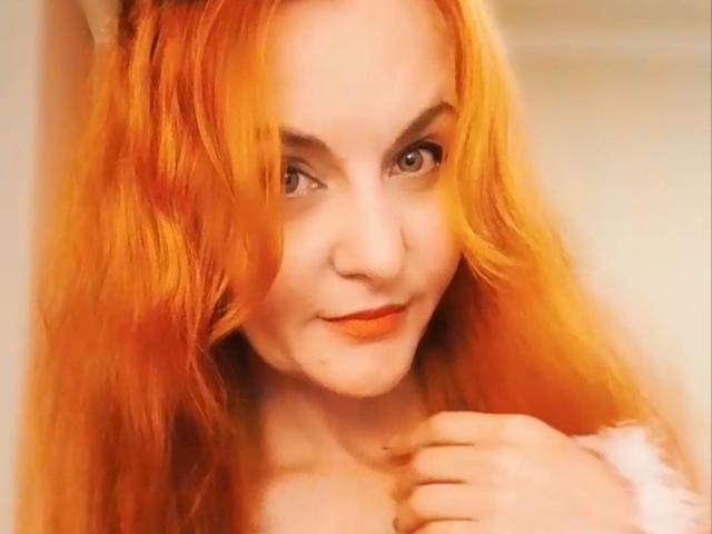 JasminsLove with fiery orange-red hair, slight smile, and light makeup, holding her chin, showing soft freckles and pale skin, in a warm-lit, slightly blurry selfie.