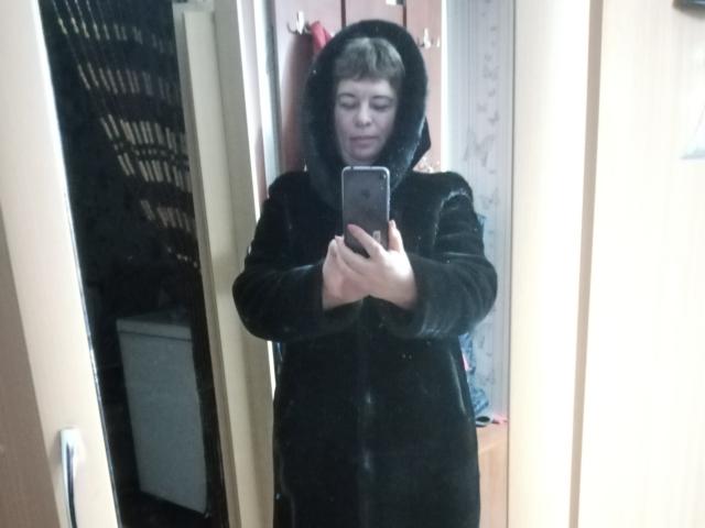 LeeNell stands in a mirror selfie, wearing a black hoodie with a hood up, holding a phone, showing a neutral expression, with short brown hair.