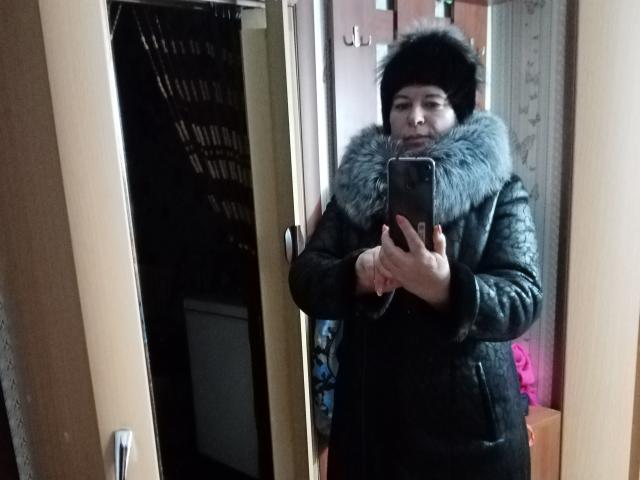LeeNell taking a mirror selfie, wearing a black fur hat, gray fur-lined coat, holding a phone, standing in a wooden room with a door and shelf in background.