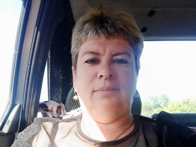 LeeNell, chubby face with short brown hair, slight smile, wearing a brown shirt, sitting in a car with a small dog on her lap, sunlight on her face.