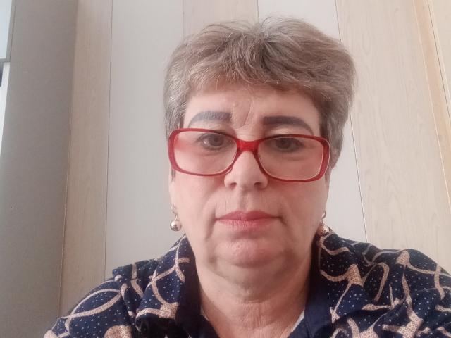 LeeNell with short gray hair, red glasses, and a patterned dark blue shirt, showing a serious face, small earrings visible, light wood background.