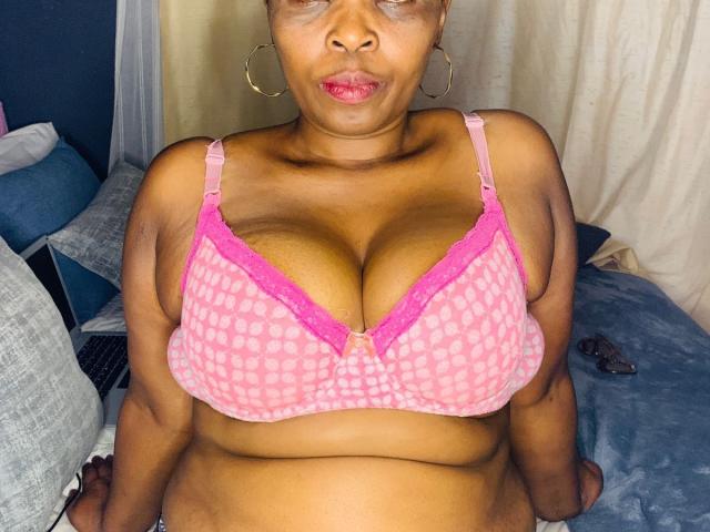 SexyRubi69, a fat brown-skinned woman with big tits in a pink checkered bra, puffy lips, and gold hoop earrings, sitting on a bed, looking up.