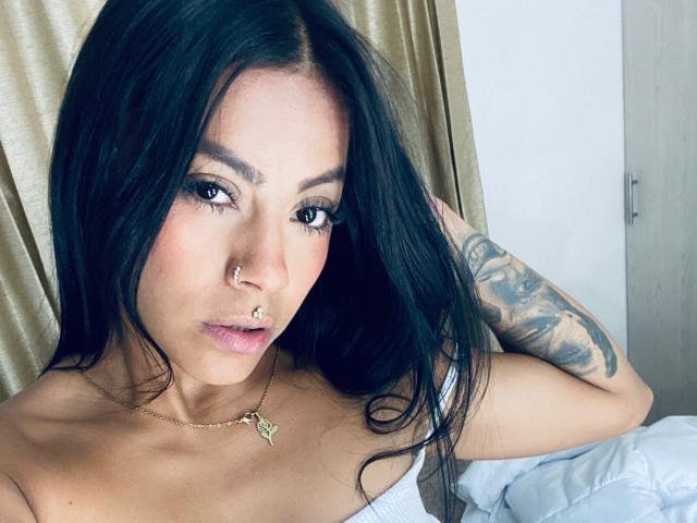 Amberv has long black hair, dark eyes, and a nose ring. She's wearing a gold necklace and has a tattoo on her right arm. She's lying in bed.