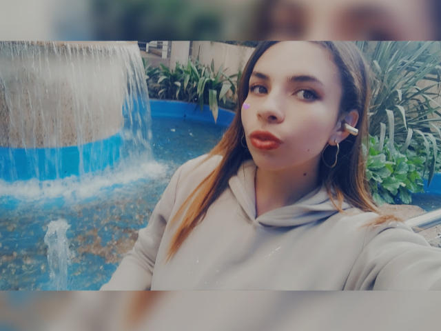 VioletaStone with pouty lips, light brown hair, wearing a gray hoodie, standing by a blue-tiled water feature, small earrings, blurred foreground.
