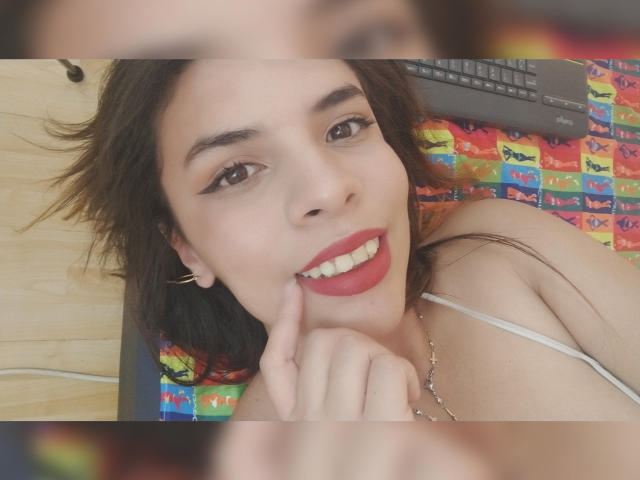 VioletaStone has dark brown hair, red lipstick, and is smiling with her finger on her chin, wearing a white spaghetti strap top, lying on a colorful quilted bed.