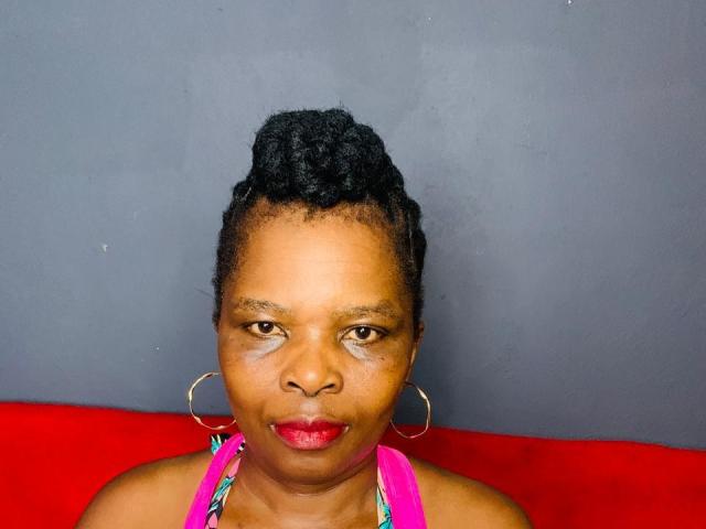 SexyRubi69 has dark skin, red lipstick, and small hoop earrings, wearing a pink halter top with colorful straps, and has her black hair in a neat bun.